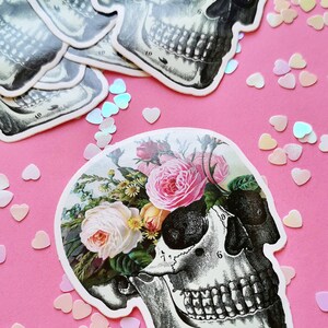 May include: A black and white anatomical skull sticker with a floral design. The skull is surrounded by pink, white, and yellow roses and other flowers. The sticker is on a pink background.