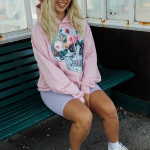 May include: A pink hoodie with a floral skull graphic and a pink fuzzy bucket hat with a red heart. The person is wearing light purple biker shorts and white sneakers with a black and white pattern.