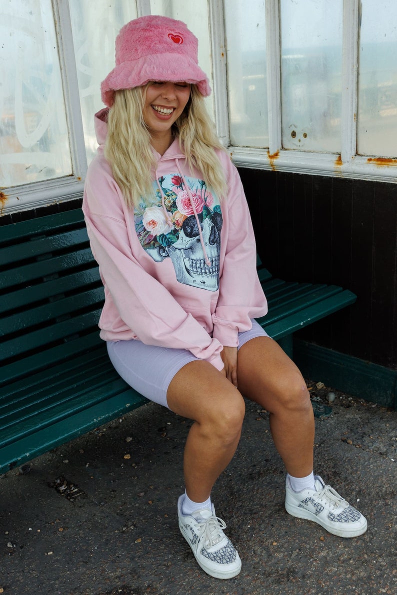 May include: A pink hoodie with a floral skull graphic and a pink fuzzy bucket hat with a red heart. The person is wearing light purple biker shorts and white sneakers with a black and white pattern.
