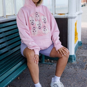 May include: A woman wearing a pink hoodie with a graphic print of multiple baby faces. She is sitting on a green bench and wearing white sneakers with a black and white pattern.