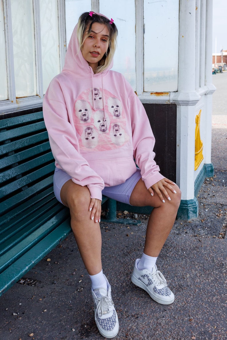 May include: A woman wearing a pink hoodie with a graphic print of multiple baby faces. She is sitting on a green bench and wearing white sneakers with a black and white pattern.