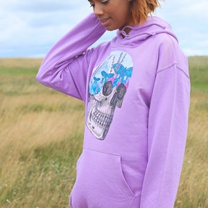 May include: A lavender hoodie with a front pocket and a drawstring hood. The graphic on the front features a detailed skull design with butterflies and floral elements in shades of blue and pink. The hoodie is made of a soft material.