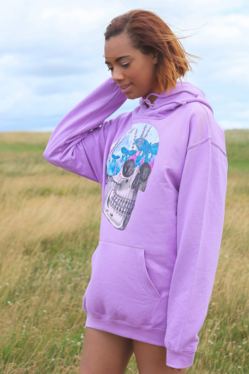 May include: A lavender hoodie with a front pocket and a drawstring hood. The graphic on the front features a detailed skull design with butterflies and floral elements in shades of blue and pink. The hoodie is made of a soft material.
