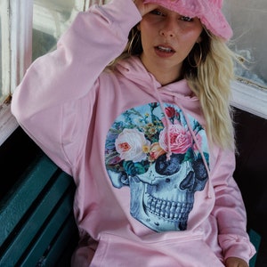 May include: A pink hoodie with a floral skull graphic print. The hoodie has a drawstring hood and a large pocket on the front. The model is wearing a pink fuzzy bucket hat with a red heart on it.