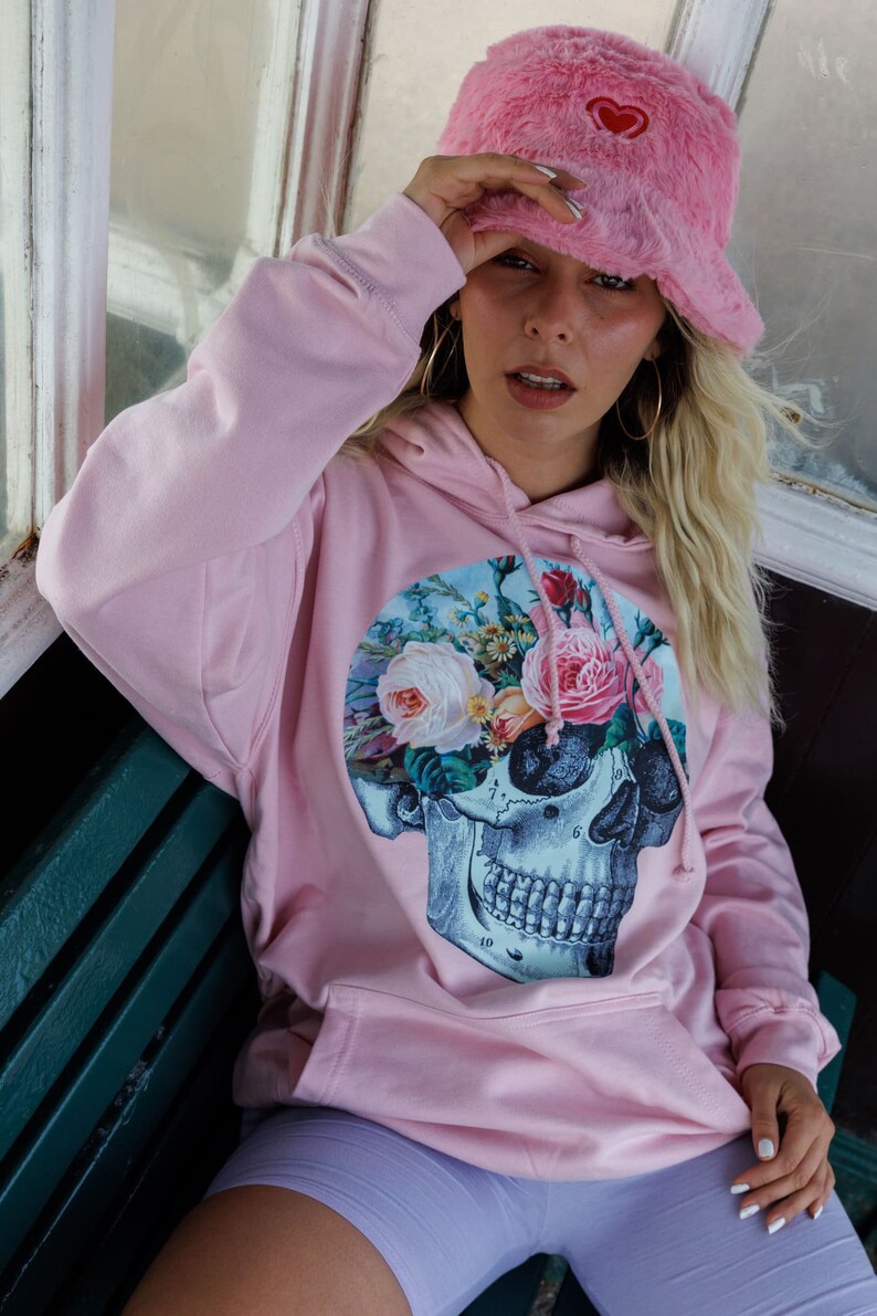 May include: A pink hoodie with a floral skull graphic print. The hoodie has a drawstring hood and a large pocket on the front. The model is wearing a pink fuzzy bucket hat with a red heart on it.
