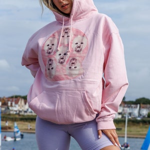 May include: A pink hoodie with a graphic print of multiple baby faces in a circle. The hoodie has a drawstring hood and a kangaroo pocket.