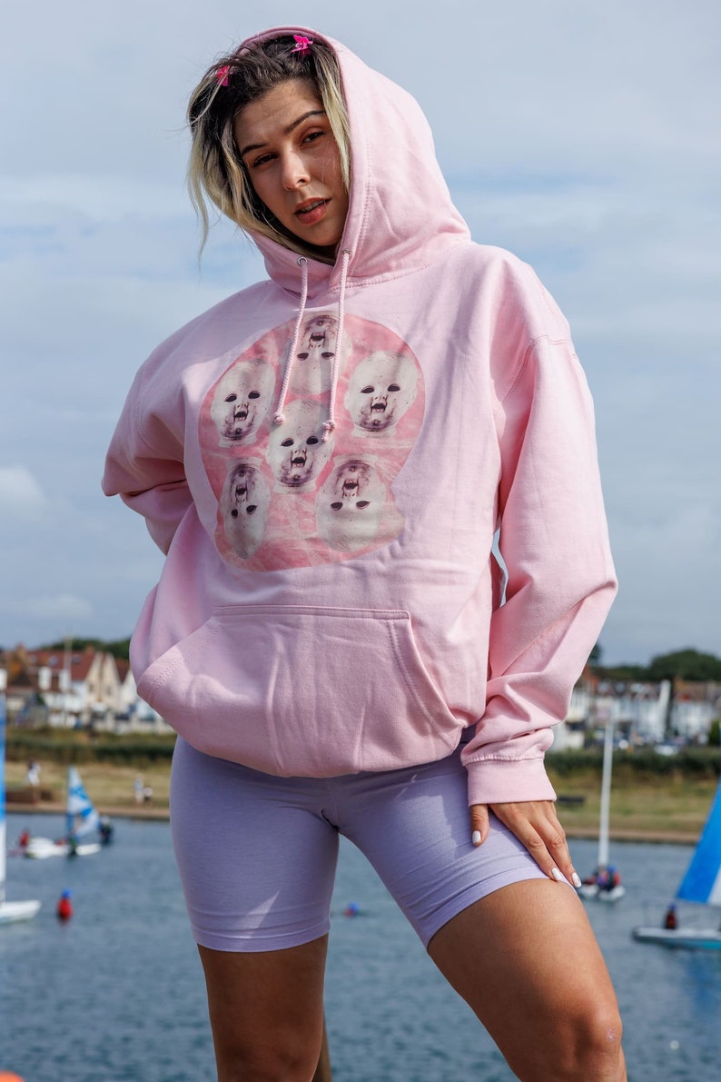 May include: A pink hoodie with a graphic print of multiple baby faces in a circle. The hoodie has a drawstring hood and a kangaroo pocket.