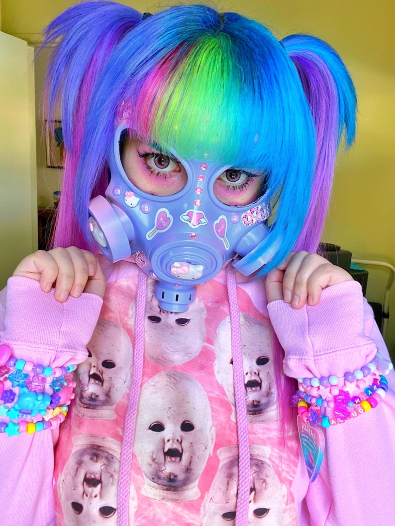 May include: A person with bright blue and pink hair wearing a pastel purple gas mask with stickers and a pink hoodie with a repeating pattern of doll faces.