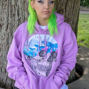 May include: A lavender hoodie featuring a skull graphic with a floral and butterfly design. The graphic is a mix of blue, pink, and white. The hoodie has a drawstring hood and a front pocket.