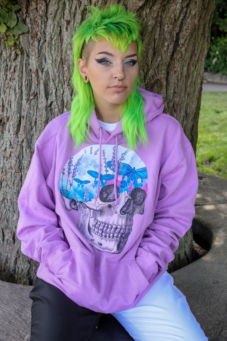 May include: A lavender hoodie featuring a skull graphic with a floral and butterfly design. The graphic is a mix of blue, pink, and white. The hoodie has a drawstring hood and a front pocket.