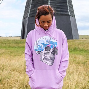 May include: A lavender hoodie with a skull graphic. The skull design features butterflies, flowers, and a moon. The hoodie has a front pocket and a drawstring hood. The background includes a windmill and a field.
