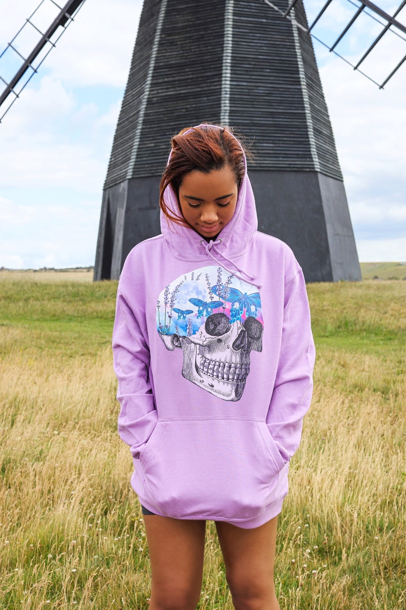 May include: A lavender hoodie with a skull graphic. The skull design features butterflies, flowers, and a moon. The hoodie has a front pocket and a drawstring hood. The background includes a windmill and a field.