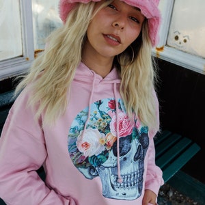 May include: A pink hoodie with a floral skull graphic. The hoodie has a drawstring hood and a kangaroo pocket. The model is wearing a pink fuzzy bucket hat.