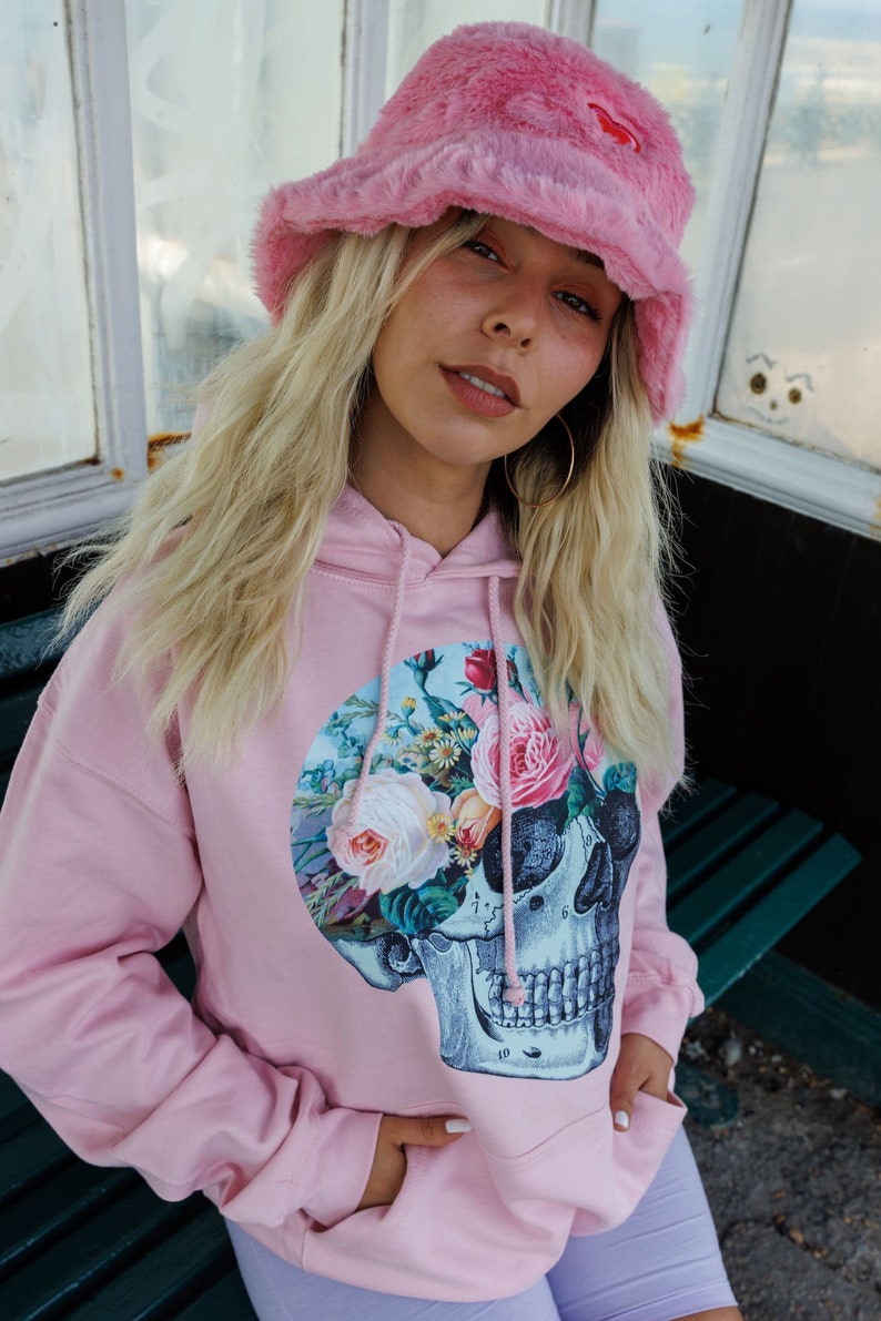 May include: A pink hoodie with a floral skull graphic. The hoodie has a drawstring hood and a kangaroo pocket. The model is wearing a pink fuzzy bucket hat.