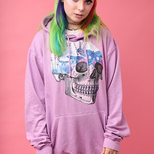 May include: A lilac-colored hoodie with a large skull graphic on the front. The skull design features a floral and butterfly motif. The model is wearing plaid pants and has rainbow-colored hair.