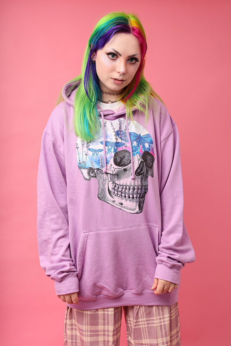 May include: A lilac-colored hoodie with a large skull graphic on the front. The skull design features a floral and butterfly motif. The model is wearing plaid pants and has rainbow-colored hair.