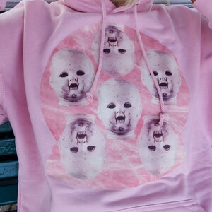 May include: Pink hoodie with a graphic print of a circle of baby doll faces with open mouths. The faces are in a pink and white marble pattern.