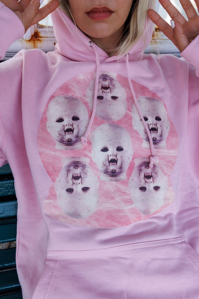 May include: Pink hoodie with a graphic print of a circle of baby doll faces with open mouths. The faces are in a pink and white marble pattern.