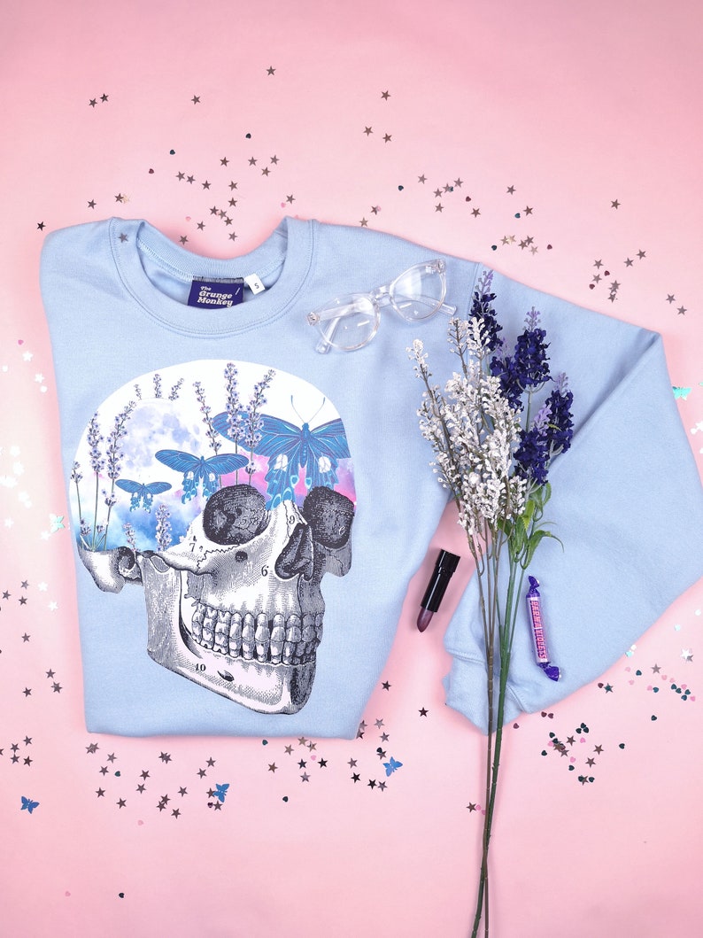 Pu&ograve; includere: Felpa blu chiaro con una stampa grafica di uno scheletro con lavanda e farfalle. Il testo "The Grunge Monkey" &egrave; sull'etichetta.