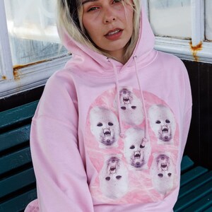 May include: A pink hoodie with a graphic print of multiple baby doll faces with open mouths. The faces are arranged in a circle on a pink background.