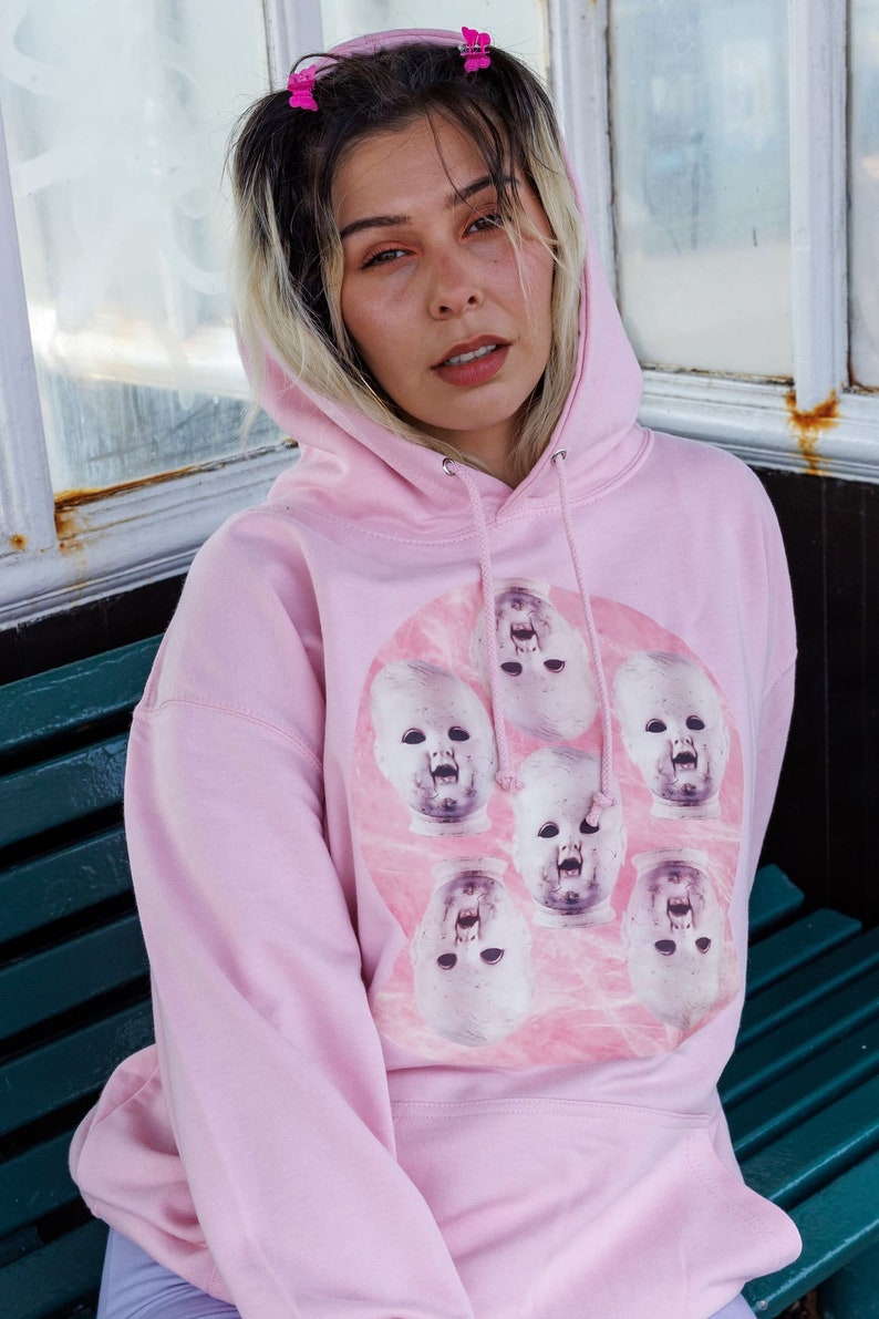 May include: A pink hoodie with a graphic print of multiple baby doll faces with open mouths. The faces are arranged in a circle on a pink background.