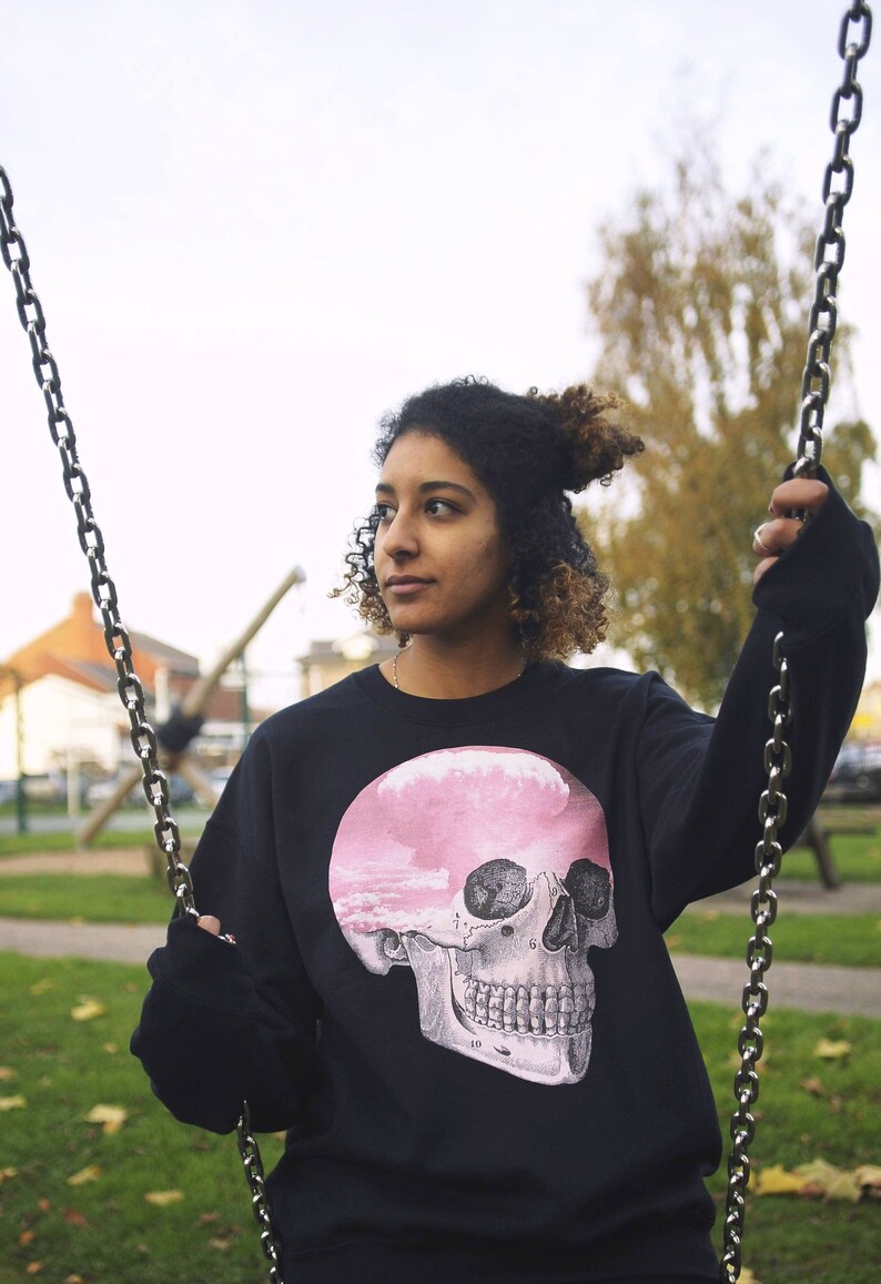 May include: A person wearing a black sweatshirt with a pink and white skull graphic. The skull has a cloudy sky inside of it. The person is sitting on a swing.