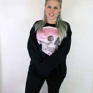 May include: A black sweatshirt featuring a pink and white skull graphic. The skull has a detailed anatomical design. The model is wearing black jeans with a distressed look.
