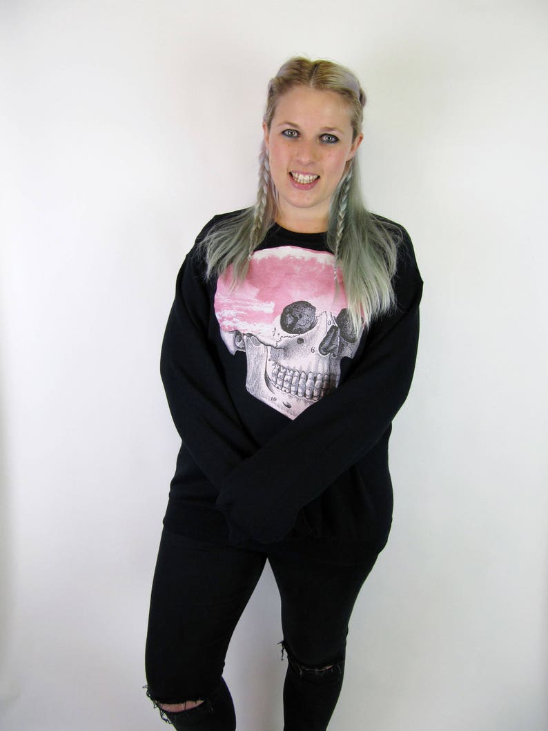 May include: A black sweatshirt featuring a pink and white skull graphic. The skull has a detailed anatomical design. The model is wearing black jeans with a distressed look.