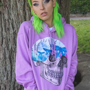 May include: A lilac hoodie featuring a skull graphic with butterflies and lavender. The skull design is detailed with anatomical features. The hoodie has a drawstring hood and a kangaroo pocket.