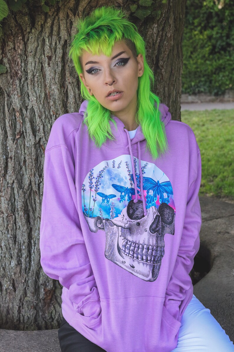 May include: A lilac hoodie featuring a skull graphic with butterflies and lavender. The skull design is detailed with anatomical features. The hoodie has a drawstring hood and a kangaroo pocket.