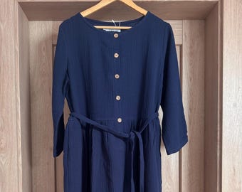 Puede incluir: Vestido midi azul marino con mangas largas, cuello redondo y abotonadura frontal. El vestido tiene un cinturón de tela a juego y está colgado de una percha de madera. El vestido está hecho de una tela texturizada.