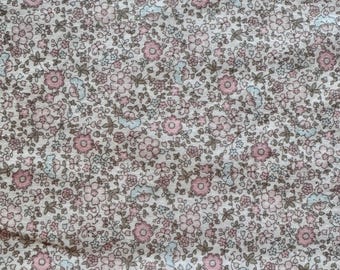 May include: Close-up of a fabric with a floral pattern. The design features small pink and light blue flowers with green leaves on a cream-colored background. The fabric has a slightly wrinkled texture.