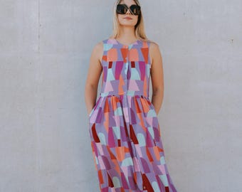 May include: A sleeveless, button-down dress with a colourful geometric pattern in shades of purple, pink, orange, and teal. The dress is knee-length and worn with black sandals and sunglasses.