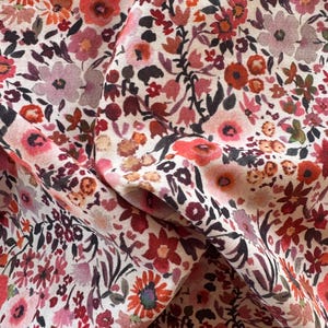May include: Close-up of floral fabric with a cream background. The fabric features a dense pattern of small flowers in shades of red, pink, and orange, with dark green and black leaves. The fabric is folded, showing texture and depth.