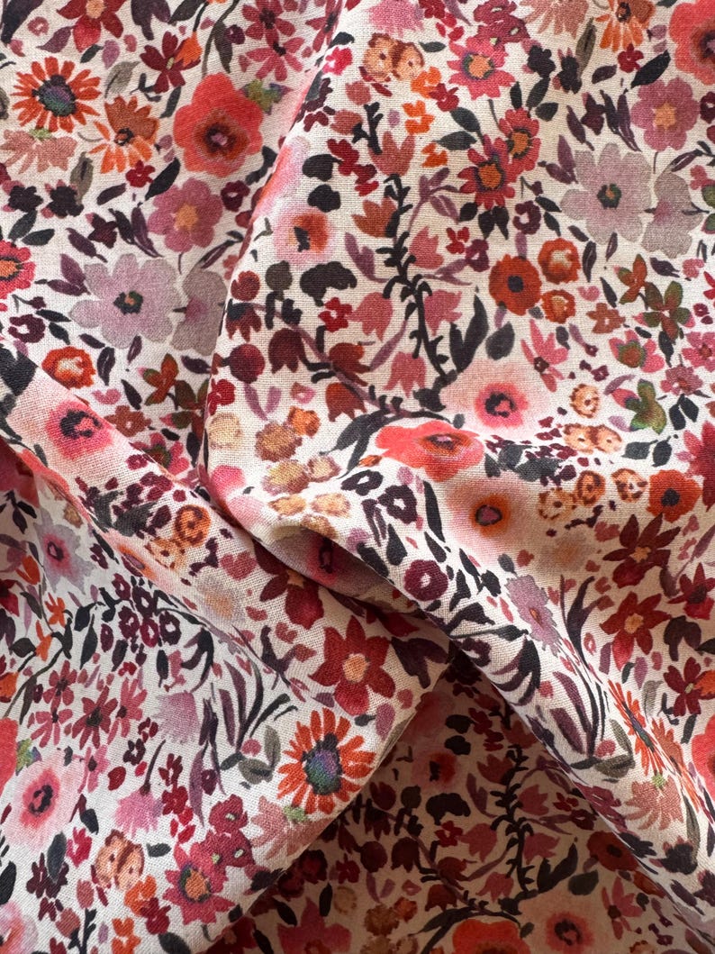 May include: Close-up of floral fabric with a cream background. The fabric features a dense pattern of small flowers in shades of red, pink, and orange, with dark green and black leaves. The fabric is folded, showing texture and depth.