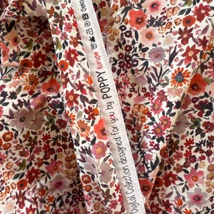 May include: Close-up of floral fabric with a cream background and a dense pattern of red, pink, and orange flowers. A white ribbon with text reading "Digital Collection designed for you by POPPY Europe" is draped across the fabric. The fabric is likely cotton or a similar material.