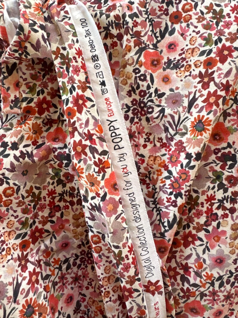 May include: Close-up of floral fabric with a cream background and a dense pattern of red, pink, and orange flowers. A white ribbon with text reading "Digital Collection designed for you by POPPY Europe" is draped across the fabric. The fabric is likely cotton or a similar material.
