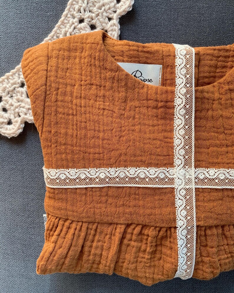 K&ouml;nnte beinhalten: Ein braunes, &auml;rmelloses Kleid mit wei&szlig;em Spitzenbesatz. Das Kleid hat einen R&uuml;schen-Saum und ein kleines wei&szlig;es Etikett mit dem Wort "Rose" darauf.