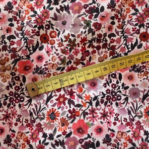 May include: Floral fabric with a dense pattern of red, pink, and orange flowers on a white background. A yellow measuring tape is laid across the fabric, showing measurements in inches and centimeters.