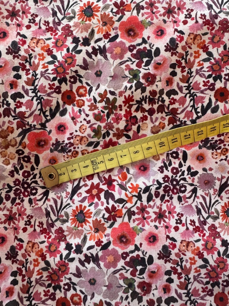 May include: Floral fabric with a dense pattern of red, pink, and orange flowers on a white background. A yellow measuring tape is laid across the fabric, showing measurements in inches and centimeters.