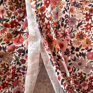 May include: Close-up of floral fabric with a cream background and a pattern of red, pink, and orange flowers. The fabric has a delicate, lightweight appearance, with a white strip of fabric visible in the center. The text on the white strip is blurry.