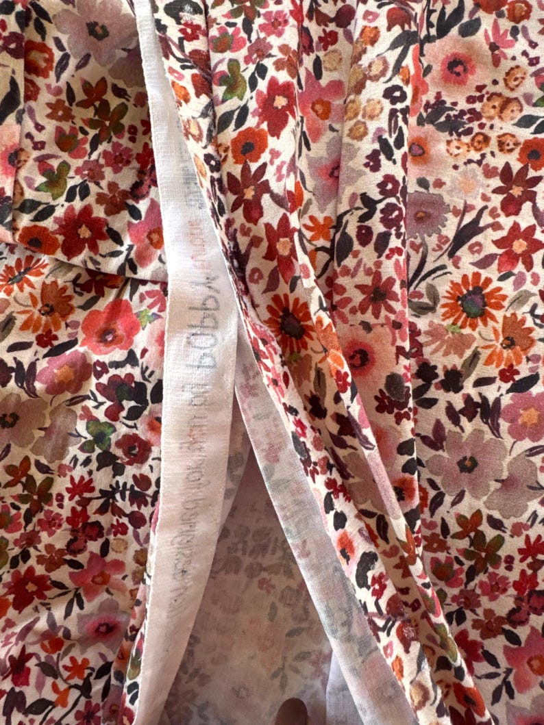 May include: Close-up of floral fabric with a cream background and a pattern of red, pink, and orange flowers. The fabric has a delicate, lightweight appearance, with a white strip of fabric visible in the center. The text on the white strip is blurry.