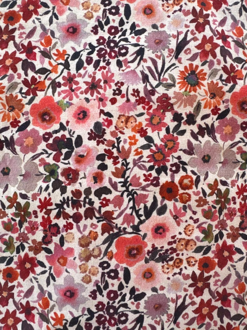 May include: A close-up of a floral fabric with a dense pattern of small flowers in shades of red, pink, orange, and purple. The flowers are set against a cream-colored background, creating a vibrant and detailed textile design.