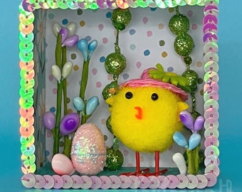 May include: A yellow chick with a pink hat and a sparkly blue egg in a colorful frame with flowers and green leaves.
