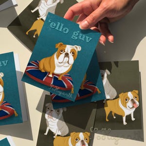 以下が含まれることがあります： ユニオンジャックのクッションに座っている漫画のブルドッグが描かれたグリーティングカード。カードはターコイズブルーの背景で、白い文字で「'ello guv」と書かれています。