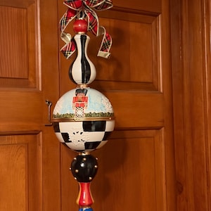 May include: A decorative hanging ornament with a whimsical design. It features a black and white checkered sphere, a blue and red base, and a red and gold plaid bow. The ornament is suspended by a gold chain.
