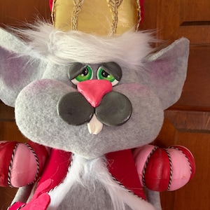 May include: A plush gray mouse wearing a red and gold crown, red sash, and pink and red decorative accents. The mouse has green eyes, a pink nose, and a white beard. The toy is a whimsical decorative item.