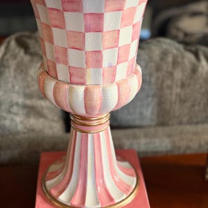 May include: A decorative urn with a pink and white checkered pattern on the upper portion. The urn has a pink and white striped base and a gold rim. The overall design is ornate and colorful.