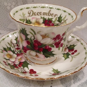 May include: A fine bone china teacup and saucer set. The design features pink flowers, red berries, and green foliage. The cup is inscribed with the word "December" in gold. The set has gold edging.