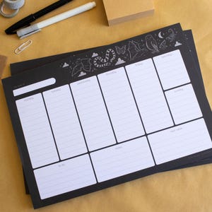May include: A black weekly planner with white sections for each day, notes, and to-do lists. The top border has a whimsical design of clouds, a snake, an owl, and other illustrations. Pens, tape, and notepads are also visible.